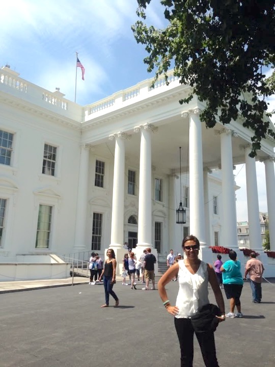 A woman wearing sunglasses stands with a hand on her hip, posing for a photo in front of the White House.