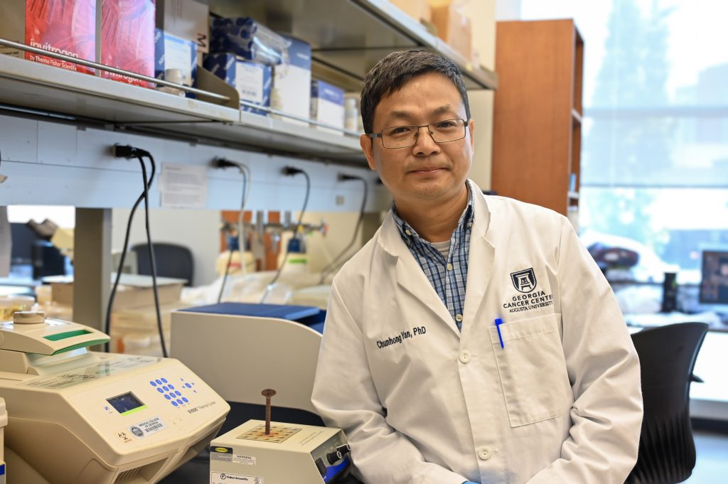 A man in a white lab coat with the words "Chunhong Yan, PhD" on the left and "Georgia Cancer Center Augusta University" on the right leans against a counter in his lab.