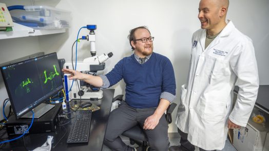 A man in a blue shirt and glasses points to a green highlight on a computer monitor, while a man in a lab coat looks on.