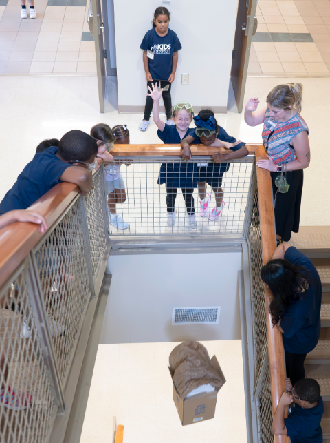 Elementary-aged children participate in a science experiment where they put padding in a box to protect an object being dropped from the top of a staircase.