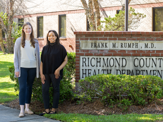 Two female college students stand next to a brick sign. The sign reads, "Richmond County Health Department."