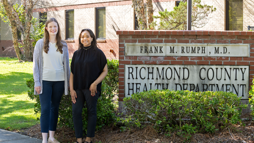 Two female college students stand next to a brick sign. The sign reads, "Richmond County Health Department."