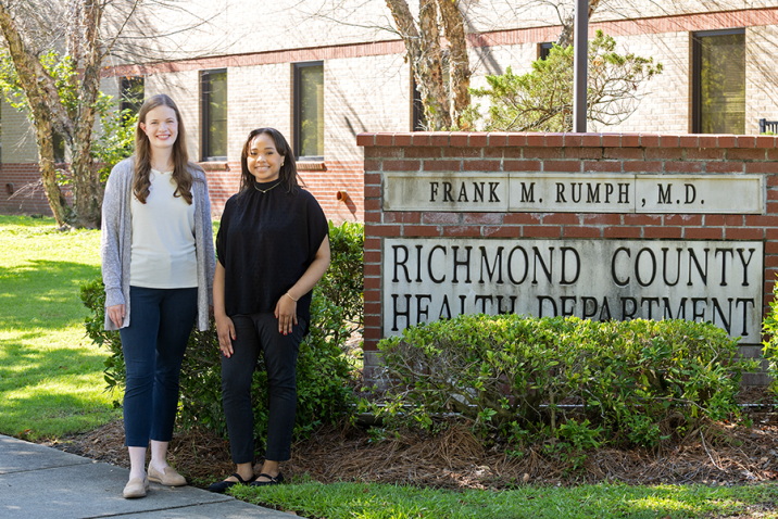 Two female college students stand next to a brick sign. The sign reads, "Richmond County Health Department."