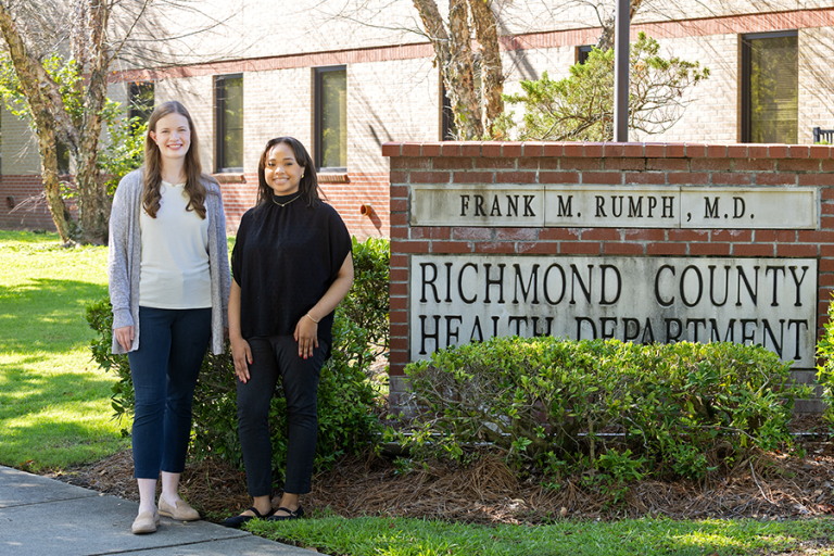 Two female college students stand next to a brick sign. The sign reads, "Richmond County Health Department."