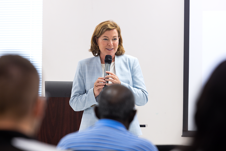 A woman holds a microphone while addressing a large crowd gathered inside a meeting room for a presentation.