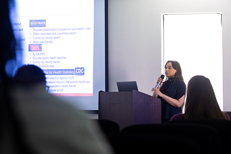 A female college student holds a microphone and stands at a podium in front of a large group of people while giving a presentation.