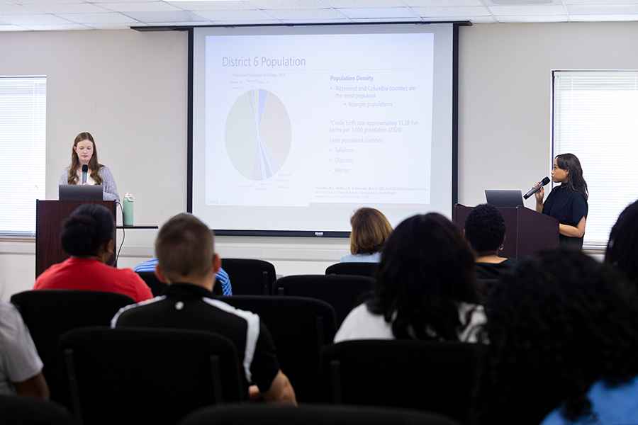 Two female college students stand at podiums at the front of a room. They are giving a presentation to a full room of people on health disparities in the state of Georgia.