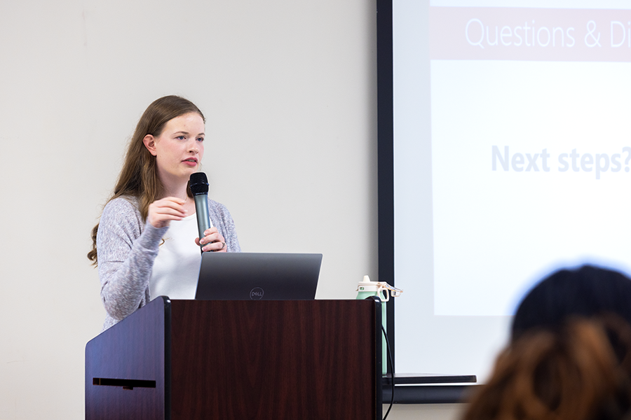 A female college student holds a microphone and stands at a podium in front of a large group of people while giving a presentation.