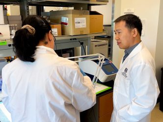 Dr. Bangxing Hong, a male, is standing in his research laboratory inside the Georgia Cancer Center's M. Bert Storey Research Building. Dr. Hong is speaking with Dr. Konstantina Kyritsi, a female scientist, about an experiment happening on a work bench in front of the two scientists.