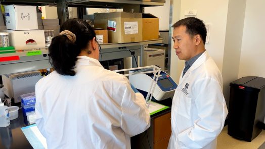 Dr. Bangxing Hong, a male, is standing in his research laboratory inside the Georgia Cancer Center's M. Bert Storey Research Building. Dr. Hong is speaking with Dr. Konstantina Kyritsi, a female scientist, about an experiment happening on a work bench in front of the two scientists.