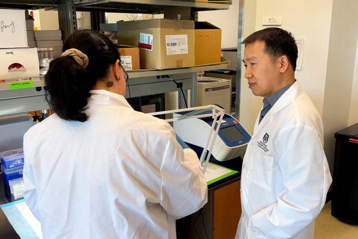 Dr. Bangxing Hong, a male, is standing in his research laboratory inside the Georgia Cancer Center's M. Bert Storey Research Building. Dr. Hong is speaking with Dr. Konstantina Kyritsi, a female scientist, about an experiment happening on a work bench in front of the two scientists.