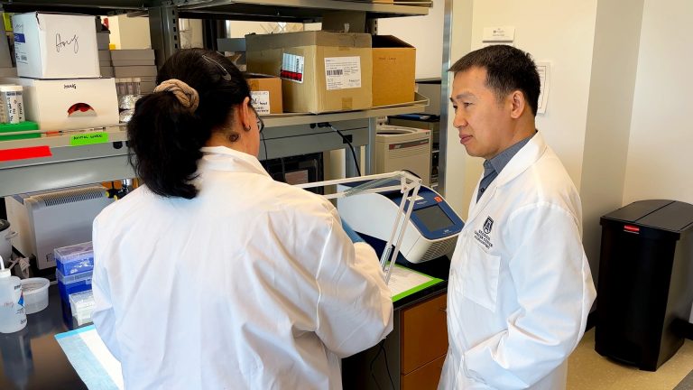 Dr. Bangxing Hong, a male, is standing in his research laboratory inside the Georgia Cancer Center's M. Bert Storey Research Building. Dr. Hong is speaking with Dr. Konstantina Kyritsi, a female scientist, about an experiment happening on a work bench in front of the two scientists.