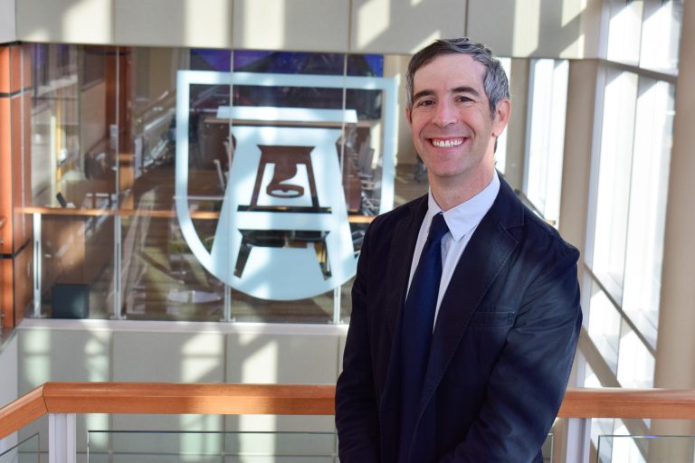 A man in a suit and tie stands in a large foyer of a college building.