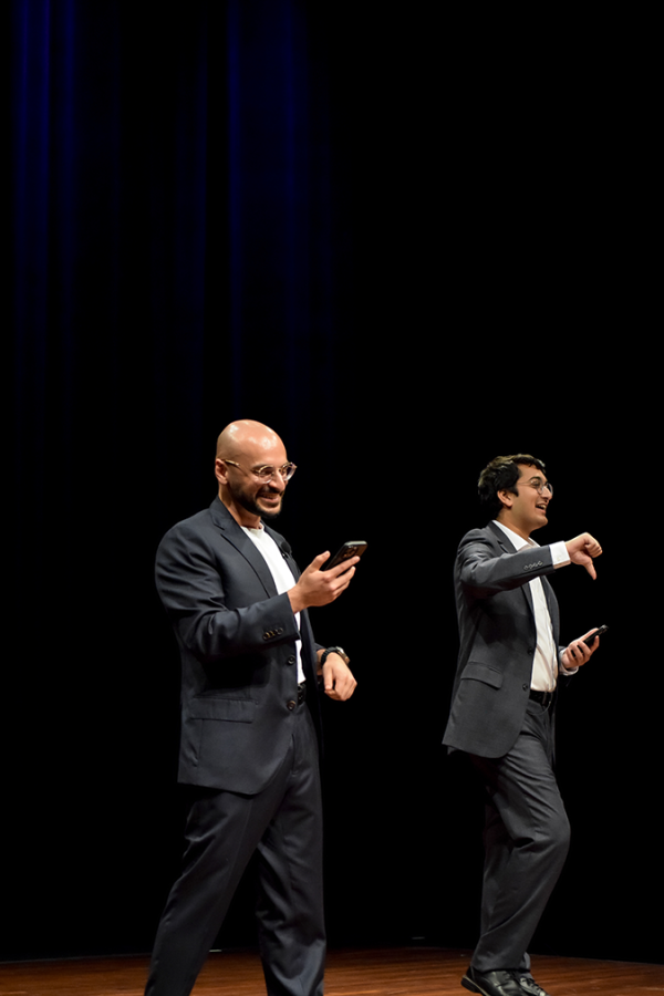Two men dressed in suits and holding their phones smile on a stage.
