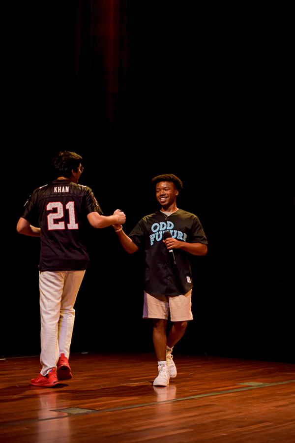 Two men holding microphones and wearing jerseys clasp hands on stage.