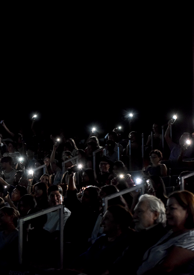 A crowd of people in a dark auditorium hold up their phone flashlights.