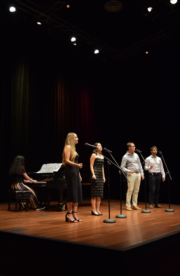 Two women and two men stand and sing into microphones on stage. A woman plays the piano behind them.