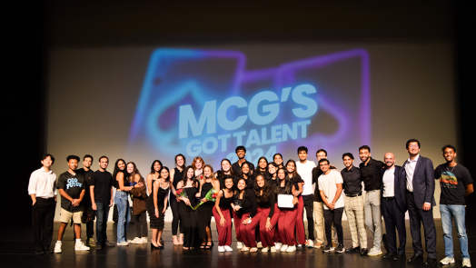 A group of medical students performing in a talent show stand in front of a backdrop that reads "MCG's Got Talent."