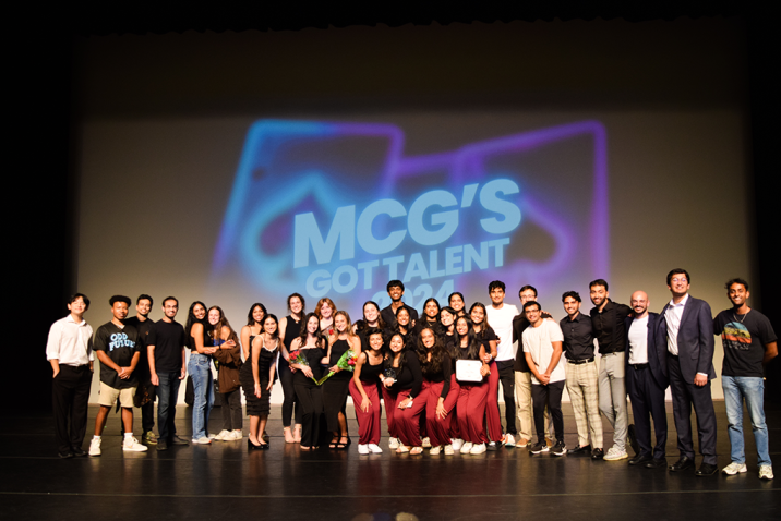 A group of medical students performing in a talent show stand in front of a backdrop that reads "MCG's Got Talent."