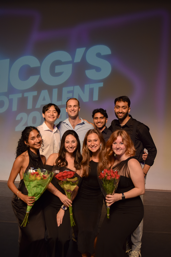 A group of men and women stand in front of a backdrop that reads "MCG's Got Talent" and smile at the camera. The women are holding flowers.