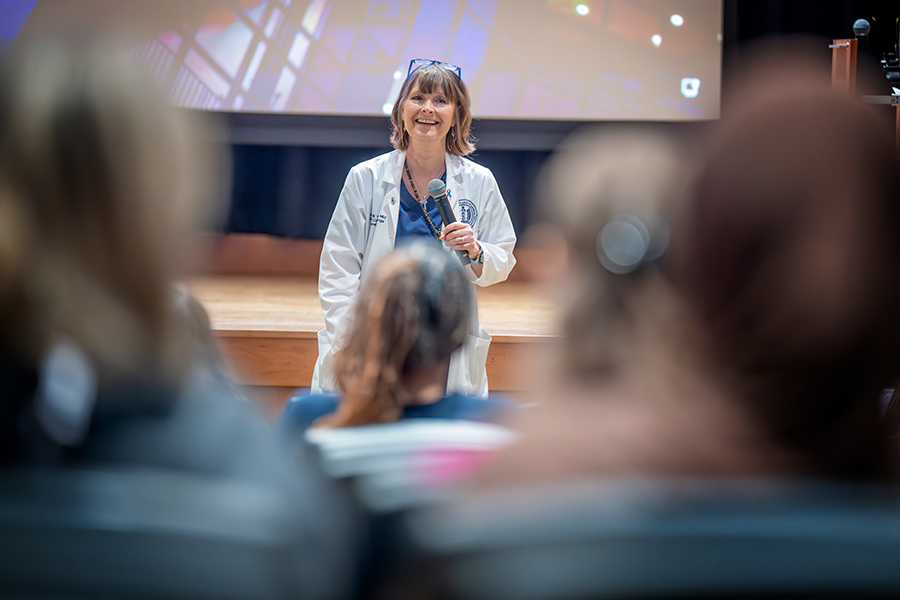 A female college professor, wearing medical scrubs and a lab coat, stands at the front of an auditorium and addresses a group of college students.