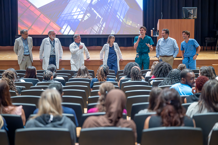 Seven college professors, including six men and one woman, stand at the front of an auditorium and speak to a large group of college students.
