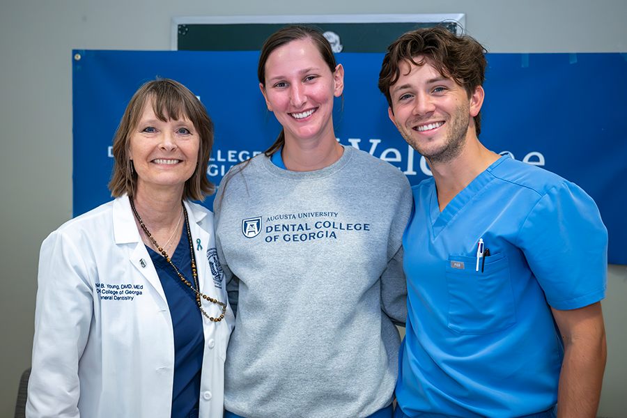 A college professor wearing medical scrubs and a lab coat stands with two students. One is wearing a sweat shirt with "Augusta University Dental College of Georgia" printed on the front.