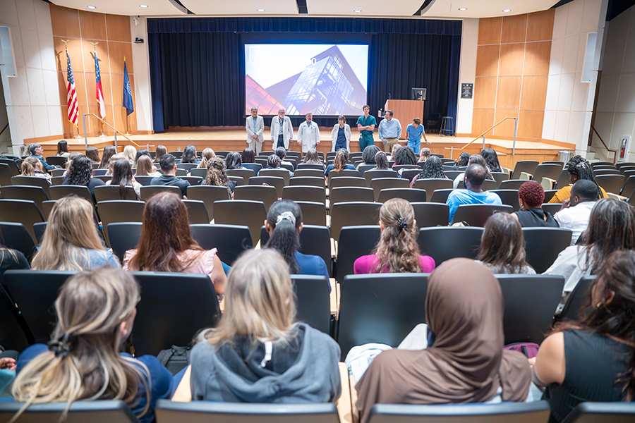 Seven college professors, including six men and one woman, stand at the front of an auditorium and speak to a large group of college students.