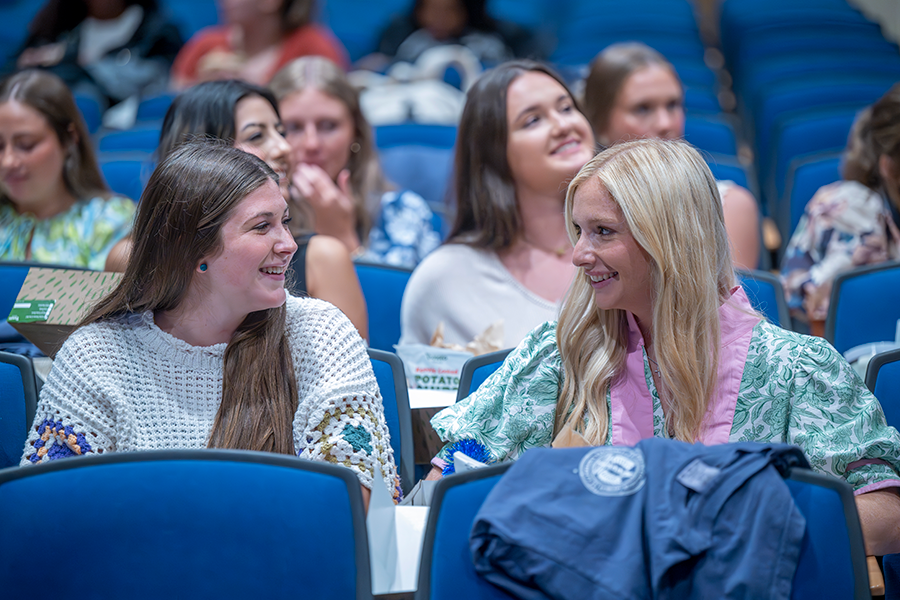 A large group of female college students sit in an auditorium, waiting for a presentation to begin.