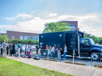 A large mobile laboratory truck parked outside of a college, with students lined up to get inside it.