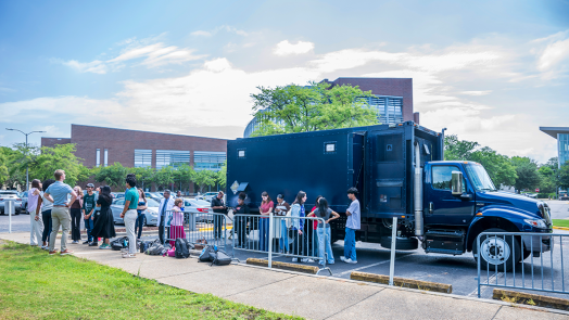 A large mobile laboratory truck parked outside of a college, with students lined up to get inside it.