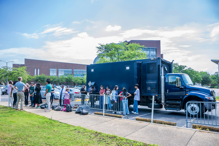 A large mobile laboratory truck parked outside of a college, with students lined up to get inside it.