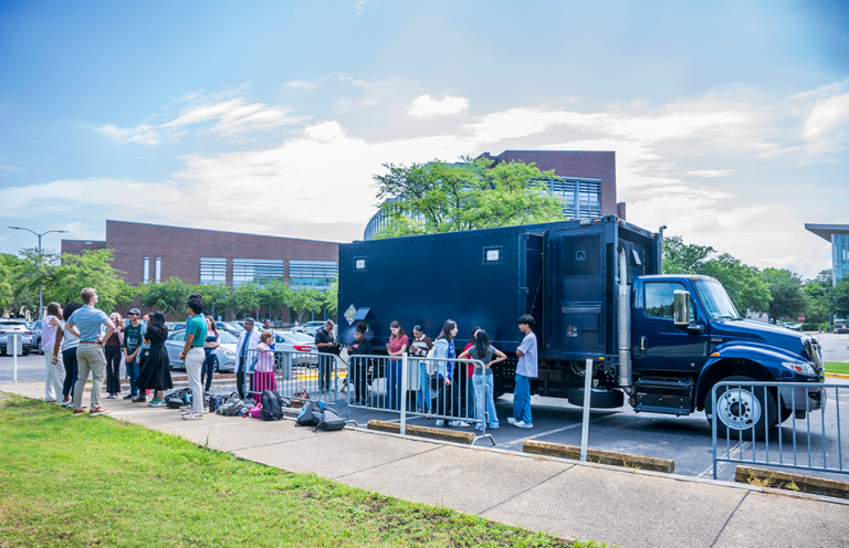 A large mobile laboratory truck parked outside of a college, with students lined up to get inside it.