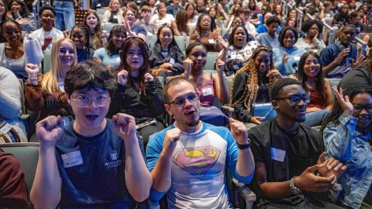 Many college-aged students since inside a building smiling and waving.