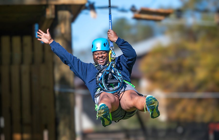 A man uses a zip line. He is wearing a harness and a helmet as he slides down towards the finish.