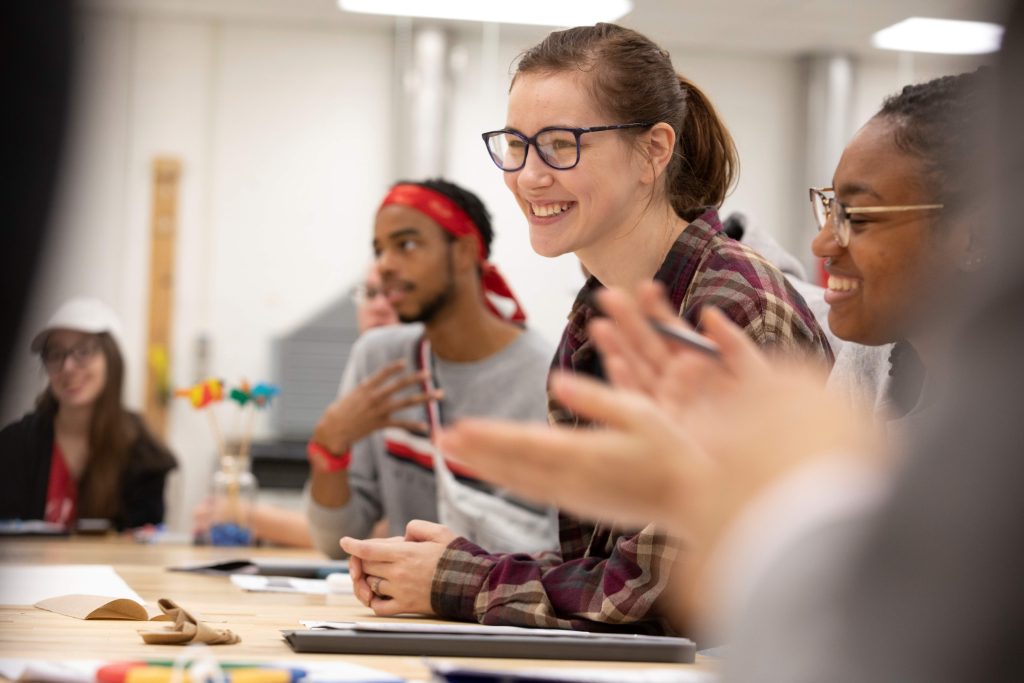 A college-aged female student sits at a classroom table smiling among other students.