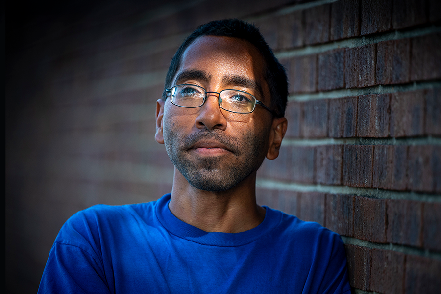 A man stands outside and leans against a brick wall.