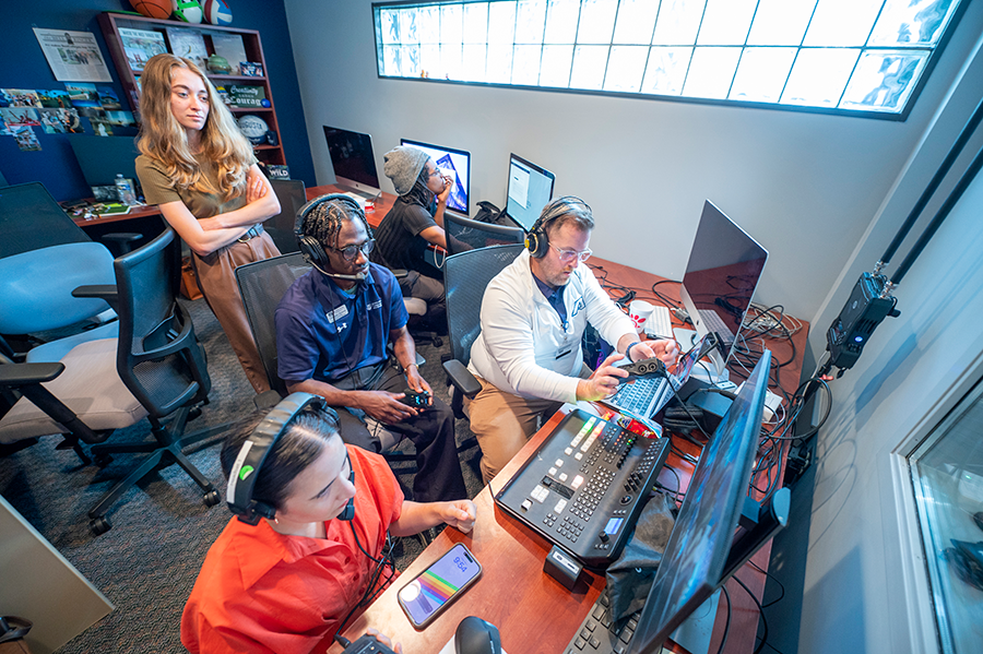 Five people, including three men and two women, sit and stand in a control room for a broadcast. They are all on headsets.