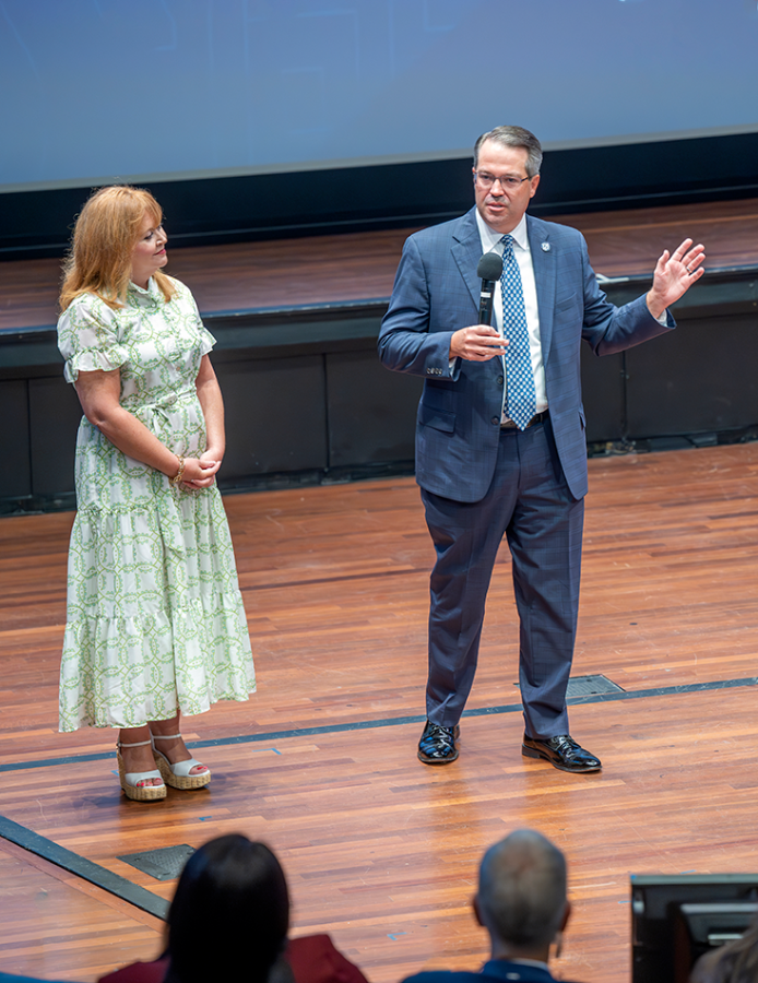 A man holding a microphone speaks on a stage with a woman standing next to him.