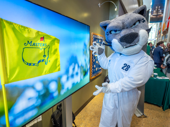 A university mascot designed after a jaguar wears a golf caddy's coveralls and points towards a screen showing a flag for the Masters golf tournament held at Augusta National Golf Club.
