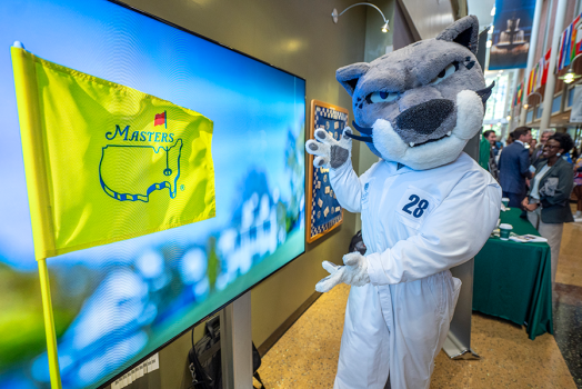 A university mascot designed after a jaguar wears a golf caddy's coveralls and points towards a screen showing a flag for the Masters golf tournament held at Augusta National Golf Club.