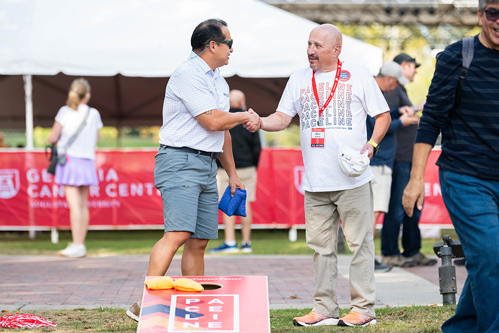 Two men shake hands behind a cornhole board at an event.