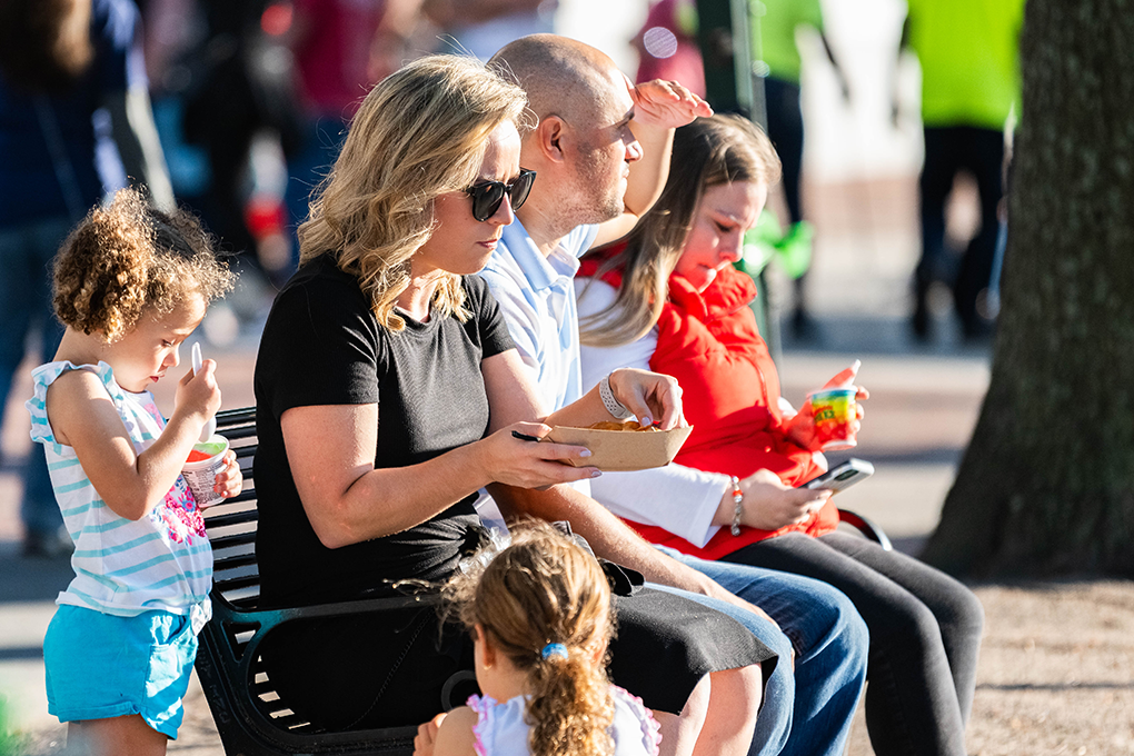 Two women and a man sit on a bench eating food at a fundraising event. Two little girls stand beside one of the women.