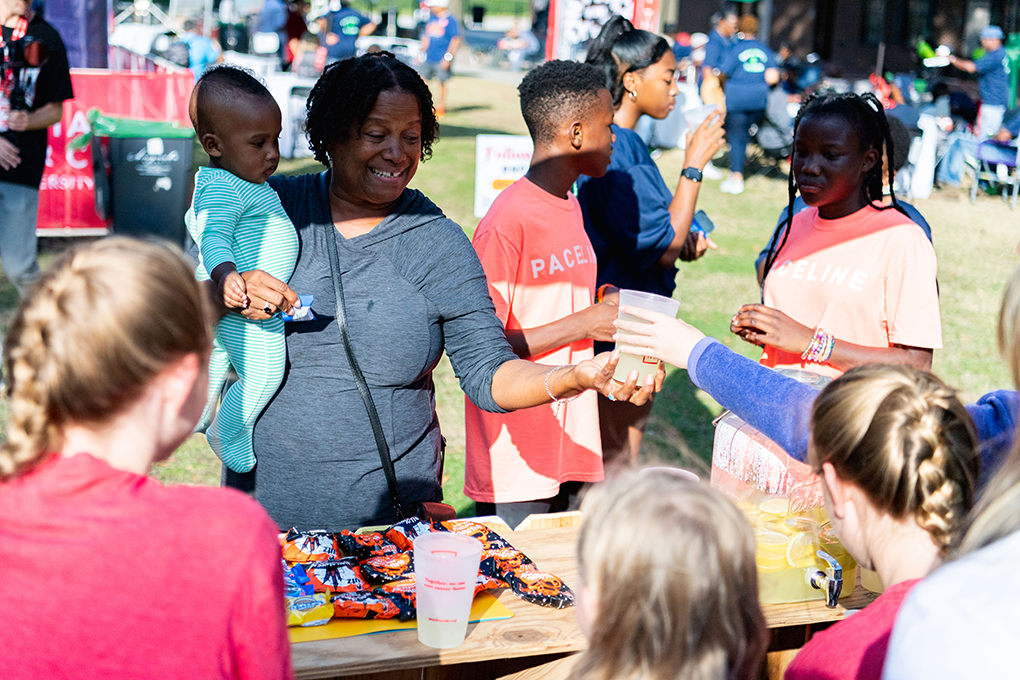 A woman holding a baby grabs a cup of lemonade from a table at an event. Two older children stand beside her.