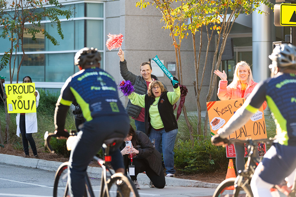 A group of people holding signs and streamers stand on the side of the road and cheer on bikers who are participating in a race.
