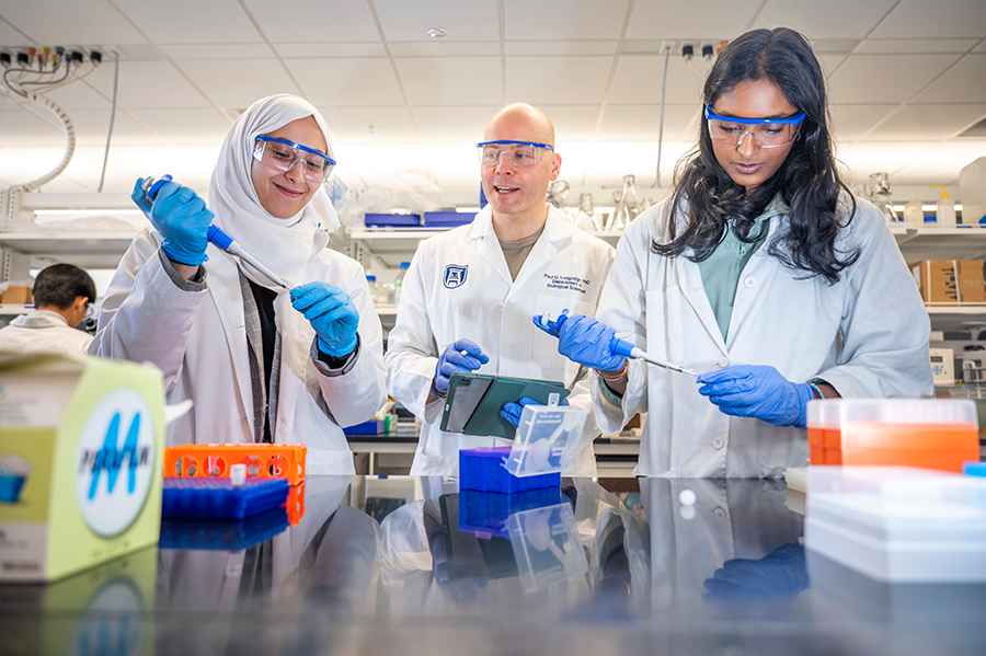A male scientist stands in between two female scientists in a lab. They are all wearing gloves, safety glasses and lab coats.