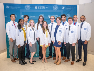 A group of 12 medical student stand in front of a backdrop following a ceremony. The backdrop has the logo and seal for the Medical College of Georgia at Augusta University on it.