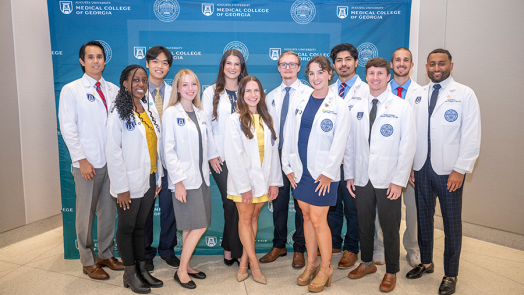 A group of 12 medical student stand in front of a backdrop following a ceremony. The backdrop has the logo and seal for the Medical College of Georgia at Augusta University on it.