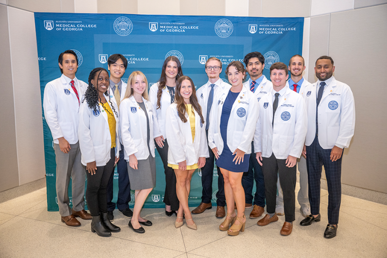 A group of 12 medical student stand in front of a backdrop following a ceremony. The backdrop has the logo and seal for the Medical College of Georgia at Augusta University on it.
