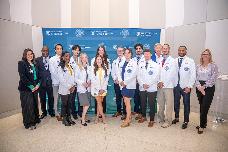 A group of 12 medical student stand in front of a backdrop following a ceremony. The backdrop has the logo and seal for the Medical College of Georgia at Augusta University on it. They are joined by four school officials.